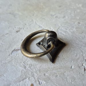 May include: Antique brass drawer pull with a diamond-shaped base and a curved handle. The hardware has a vintage aesthetic, perfect for adding a touch of classic style to furniture or cabinetry. The pull is made of metal.