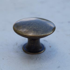 May include: A bronze cabinet knob with a rounded top and a short, cylindrical base. The knob has a slightly aged appearance, with variations in colour across its surface. The hardware is designed for use on drawers or cabinet doors.
