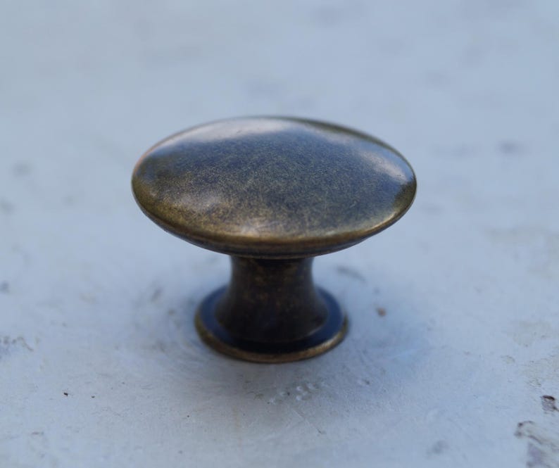 May include: A bronze cabinet knob with a rounded top and a short, cylindrical base. The knob has a slightly aged appearance, with variations in colour across its surface. The hardware is designed for use on drawers or cabinet doors.