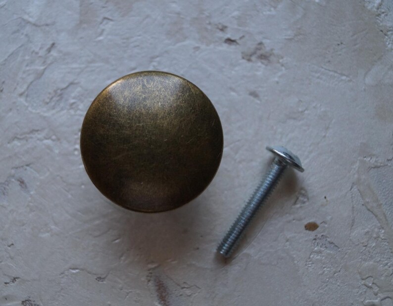 May include: A round, antique brass cabinet knob with a matching screw. The knob has a slightly domed shape and a textured surface. The screw is silver-coloured with a flat head.