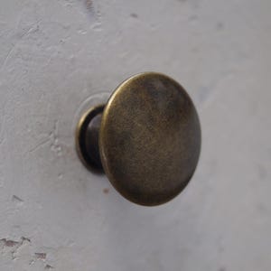 May include: Close-up of a bronze cabinet knob with a rounded, flat face. The knob is mounted on a white surface with visible texture and slight imperfections. The hardware has a vintage or antique aesthetic.