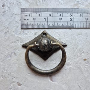 May include: Antique bronze drawer pull with a diamond-shaped backplate and a circular handle. The pull measures approximately 1 inch in width. The hardware has a vintage aesthetic, suitable for cabinets, drawers, or furniture restoration projects.