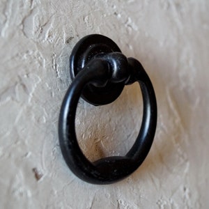 May include: A black metal ring pull door handle with a circular backplate. The handle has a rounded ring shape and is mounted on a textured, off-white surface. The hardware is likely for a cabinet or drawer.