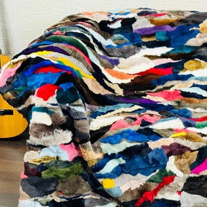 May include: A colorful patchwork blanket draped over a dark brown leather couch. The blanket features a variety of colors including blue, red, pink, and yellow. A wooden acoustic guitar is visible in the background.