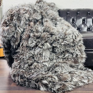 May include: A large, faux fur throw blanket in shades of gray and brown is draped over a black leather sofa. A wooden acoustic guitar leans against a white brick wall, adding a touch of rustic charm to the scene.