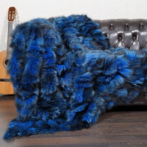 May include: A luxurious blue faux fur throw blanket draped over a dark brown leather sofa. The blanket has a plush, textured appearance with varying shades of blue and black. A wooden acoustic guitar leans against the wall in the background.