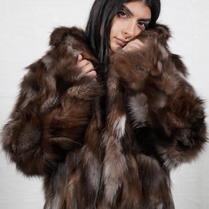 Può includere: Un cappotto di pelliccia marrone dall'aspetto strutturato. Il cappotto ha un design con cappuccio e un colore marrone scuro con accenti marrone chiaro e bianchi. Il cappotto è indossato sopra pantaloni colorati.