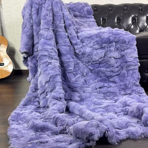 May include: A large, plush, lavender-colored throw blanket draped over a black leather sofa. The blanket has a textured, faux fur appearance. A wooden acoustic guitar leans against a white brick wall in the background.