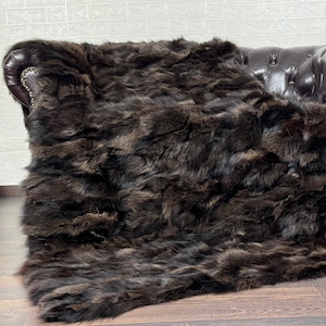 May include: A dark brown fur throw blanket draped over a brown leather Chesterfield sofa. A wooden acoustic guitar leans against a white brick wall on the left side of the image. The blanket has a shaggy texture.
