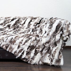 May include: A faux fur throw blanket in shades of white, gray, and brown is draped over a dark brown leather sofa. A wooden acoustic guitar leans against a white wall, adding a touch of musical charm to the scene.