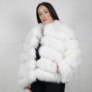 May include: A white fur jacket with a layered design. The jacket is worn over a black top and paired with black trousers. The model is standing in front of a white brick wall.