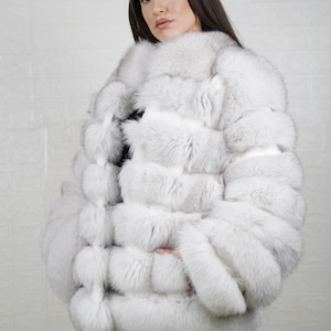 May include: A luxurious white fur coat with a textured, layered design. The coat is a light, neutral color and appears soft and plush. The garment is worn over a black top and pants, creating a striking contrast. The coat's design suggests a high-end fashion item.