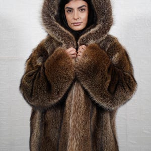 May include: A brown fur coat with a large hood. The coat has a rich, textured appearance with varying shades of brown. The hood frames the face, and the coat appears warm and luxurious. The person is wearing a black top.