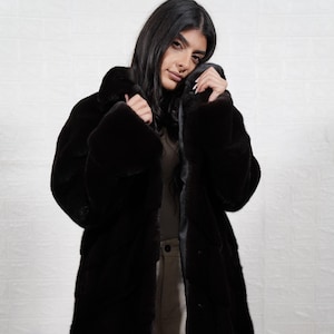 May include: A long, black faux fur coat with a notched collar. The coat features a textured, vertical design and is knee-length. The model is wearing a light-coloured shirt and trousers. The coat is a stylish outerwear option.