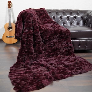 May include: A plush, burgundy-colored throw blanket draped over a dark brown leather sofa. A wooden acoustic guitar leans against the wall in the background. The blanket has a textured, ruffled appearance.
