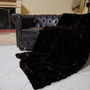 Puede incluir: Una manta de piel sintética marrón oscuro sobre un sofá Chesterfield de cuero marrón oscuro. La manta es suave y texturizada, con un color rico y profundo. El sofá está frente a una chimenea.