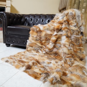 May include: A luxurious, faux fur throw blanket in shades of brown, tan, and cream, draped over a dark brown leather sofa. The blanket's texture is visible, and it rests on a tiled floor. A fireplace is in the background.