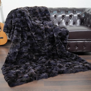 May include: A dark gray faux fur throw blanket draped over a brown leather sofa. The plush blanket has a textured surface and is spread across a wooden floor. A wooden acoustic guitar leans against a white wall in the background.