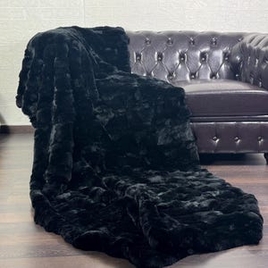 May include: A black faux fur throw blanket draped over a brown leather Chesterfield sofa. A wooden acoustic guitar leans against a white brick wall in the background. The blanket has a plush, textured appearance.