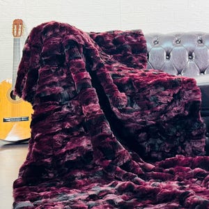 May include: A plush, burgundy faux fur throw blanket draped over a dark leather sofa. The blanket has a textured, layered appearance with black accents. A wooden acoustic guitar leans against a white brick wall.