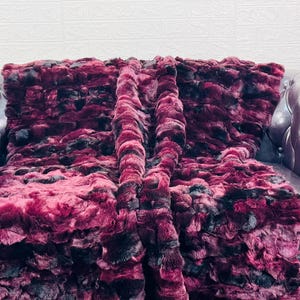 May include: A plush, faux fur throw blanket in shades of burgundy and black, draped over a burgundy leather Chesterfield sofa. The blanket's texture is dense and rippled, creating a luxurious appearance. The sofa has a button-tufted back and armrests.