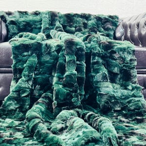 May include: A luxurious, emerald green faux fur throw blanket draped over a dark leather sofa. The blanket has a plush, textured appearance with varying shades of green and black, creating a rich, dimensional look. The sofa has a classic, tufted design.