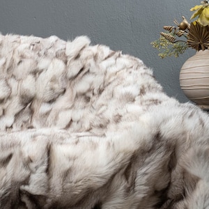 May include: A luxurious, plush throw blanket with a faux fur design. The blanket features a mix of white and gray tones, creating a textured appearance. The soft, fluffy material is draped over a surface, suggesting comfort and warmth.