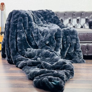 May include: A plush, dark gray faux fur throw blanket draped over a brown leather sofa. The blanket has a textured, rippled appearance. A wooden acoustic guitar leans against a white wall in the background.