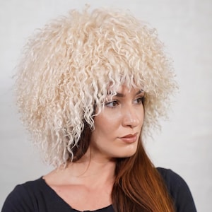 May include: A fluffy, cream-colored hat made of curly, textured material. The hat has a voluminous, rounded shape that sits atop the head, featuring dense, spiraled curls that frame the face with a soft, shaggy appearance.