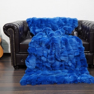 May include: A vibrant blue faux fur throw blanket draped over a dark brown leather armchair. A wooden acoustic guitar leans against the wall to the left. The blanket's texture is visible.