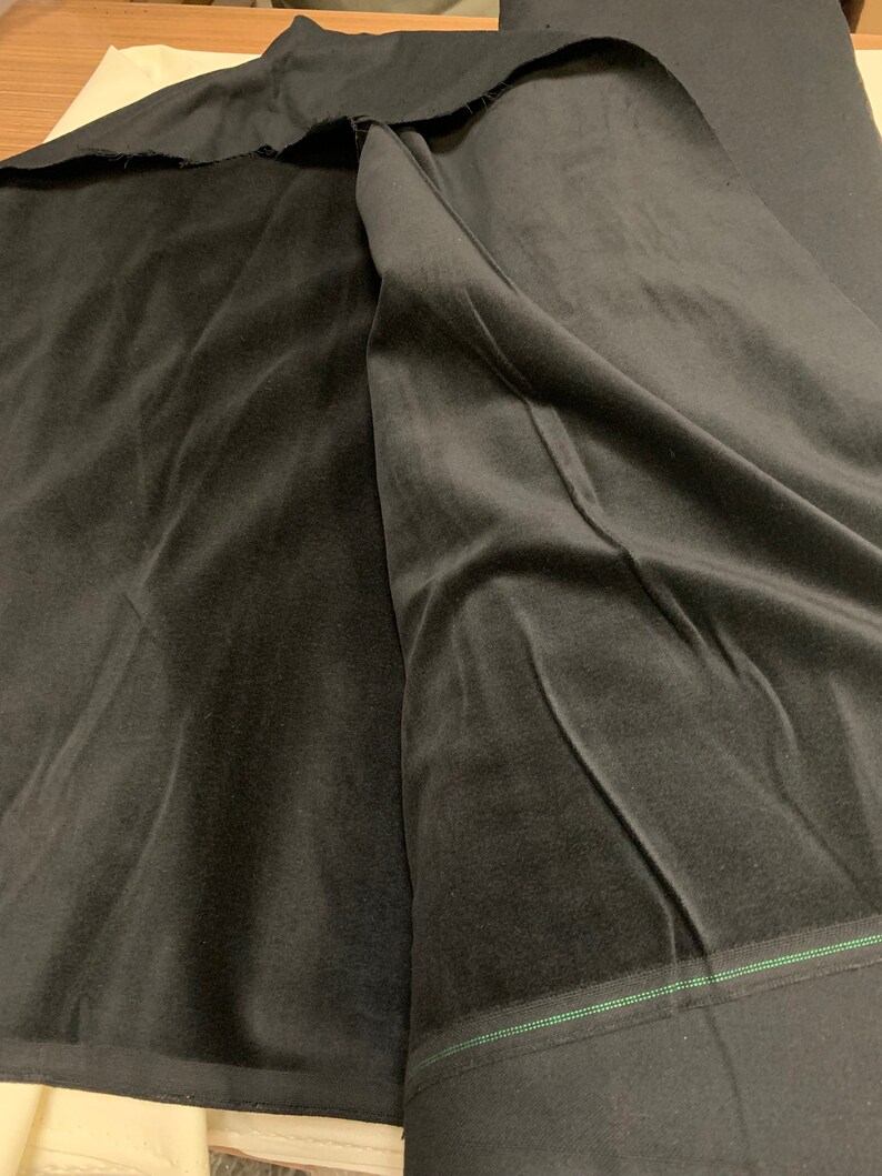 May include: A close-up of black velvet fabric, folded and draped. The fabric has a soft, plush texture and a deep, rich color. The edges of the fabric are visible, showing the cut and the backing. The fabric is likely used for clothing or home decor.