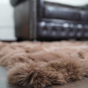 May include: A plush, brown faux fur rug with a soft, textured surface. The rug is placed on a dark wooden floor, with a blurred background featuring a guitar and a leather sofa.