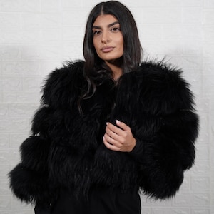 May include: A black faux fur jacket with a layered design. The jacket has a fluffy texture and a cropped length. The model is wearing a black long-sleeve top and black pants. The background is a white brick wall.