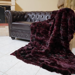 Puede incluir: Una lujosa manta de piel sintética burdeos sobre un sofá Chesterfield de cuero marrón oscuro. La manta tiene un aspecto texturizado, con toques negros. El sofá está acolchado y tiene detalles de botones.