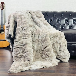 May include: A plush, faux fur blanket in shades of gray and beige is draped over a black leather sofa. A classical guitar leans against the wall in the background. The blanket's texture is visible, suggesting a soft and cozy feel.