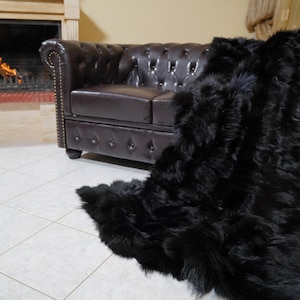 May include: A black faux fur throw draped over a brown leather Chesterfield sofa. The throw has a long, shaggy texture. A fireplace with a burning fire is visible in the background, creating a cozy atmosphere.