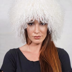 Può includere: Un cappello bianco soffice realizzato in materiale lungo, riccio e testurizzato. Il cappello ha una forma voluminosa e arrotondata che si posiziona in alto sulla testa, con fibre dense e arruffate che creano un aspetto morbido e stratificato.