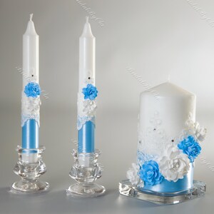 May include: A set of three decorative candles. Two tall, white candles with blue bases and floral accents sit in clear glass holders. A larger pillar candle with blue and white floral decorations rests on a square glass base. The candles are decorated with lace and rhinestones.