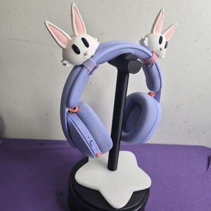 May include: Light purple over-ear headphones featuring two white rabbit head attachments with pink inner ears on the headband. The headphones are displayed on a black stand with a white star-shaped base, showcasing a textured fabric finish.