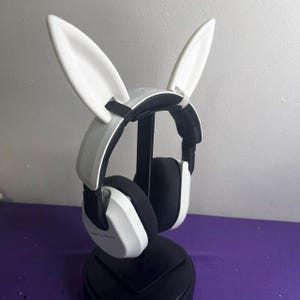 Könnte beinhalten: Weiß-schwarzer Gaming-Kopfhörer mit angebrachten weißen Hasenohren. Der Kopfhörer steht auf einem schwarzen Ständer. Die Ohrmuscheln sind schwarz und weiß, und das Kopfband ist weiß mit schwarzen Akzenten. Die Hasenohren sind groß und weiß.