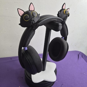 Peut inclure: Un support de casque noir orné de deux têtes de chat stylisées avec des oreilles intérieures roses et des yeux jaunes. Le support possède une base blanche en forme d'étoile à quatre branches et un montant central. Un casque noir est posé dessus.