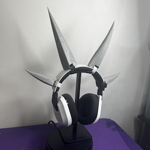 May include: White and black headphones with a unique design featuring four gray, pointed extensions. The headphones are displayed on a black stand, set against a neutral background. The overall aesthetic is modern and futuristic.