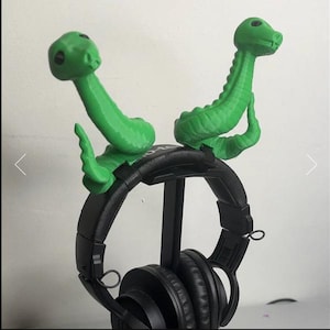 Peut inclure: Une paire d'accessoires en forme de serpent vert vif imprimés en 3D, fixés sur le dessus d'un casque audio noir. Chaque figurine de serpent présente un corps texturé et enroulé avec une tête relevée aux yeux noirs, conçue pour se clipser.