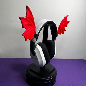 Peut inclure: Casque d'écoute blanc avec des ailes de démon rouges attachées sur les côtés. Le casque est sur un support noir.