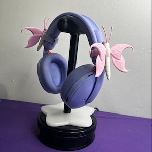 Peut inclure: Casque audio violet clair doté de deux accessoires décoratifs en forme de papillon rose sur l'arceau. Le casque est monté sur un support noir avec une base blanche en forme d'étoile, offrant un présentoir stable pour l'appareil audio.