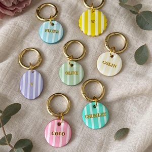 May include: A collection of seven circular and scalloped acrylic keychains with gold-tone metal rings. Each keychain features vertical stripes in pastel colors including blue, yellow, purple, green, cream, pink, and teal, with names or letters printed in gold.