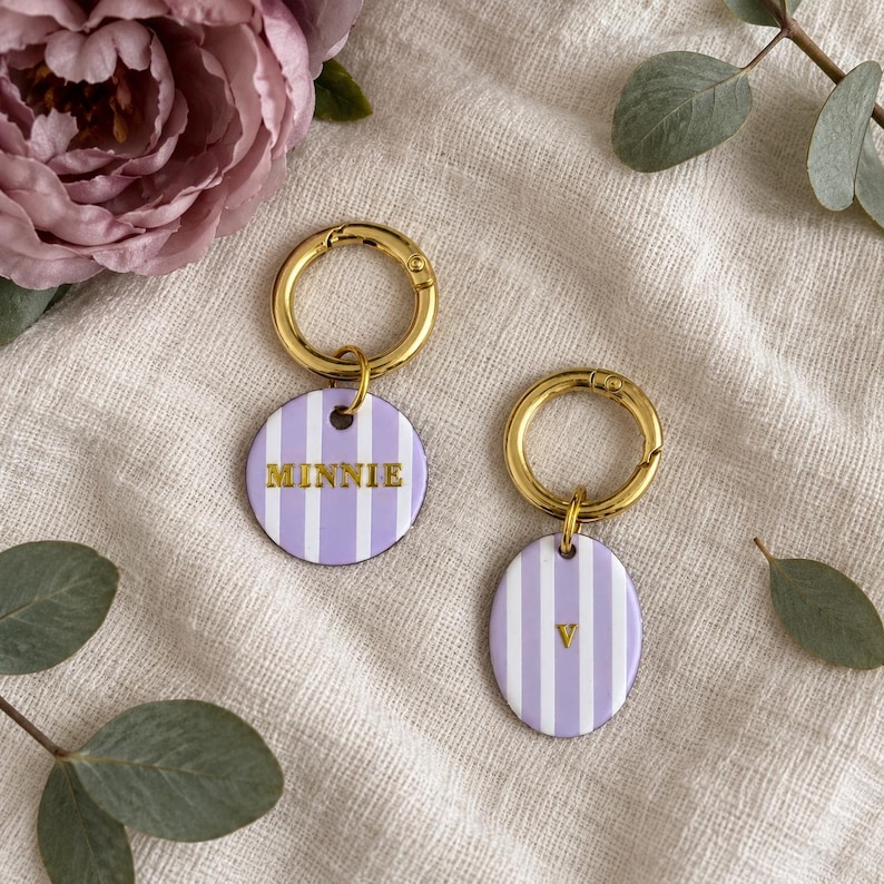 May include: Two gold-toned metal keychains featuring circular and oval pendants with a light purple and white vertical striped pattern. One pendant displays the name MINNIE in gold lettering, while the other features the letter V.