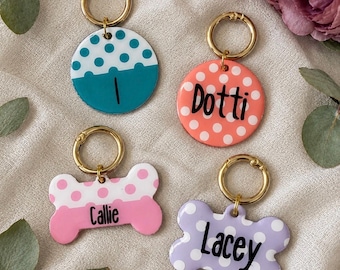 May include: Four pet ID tags with gold-tone rings. Two are circular with teal and coral polka dots, featuring the names I and Dotti. Two are bone-shaped with pink and lavender polka dots, featuring the names Callie and Lacey in black script.