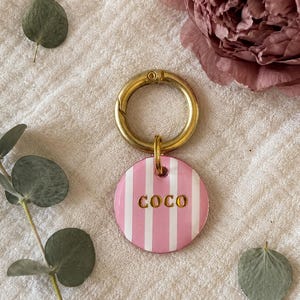 May include: A round pet tag featuring vertical pink and white stripes with the name COCO embossed in gold lettering. The tag is attached to a circular gold-toned metal keyring, providing a secure attachment point for a collar or keychain.