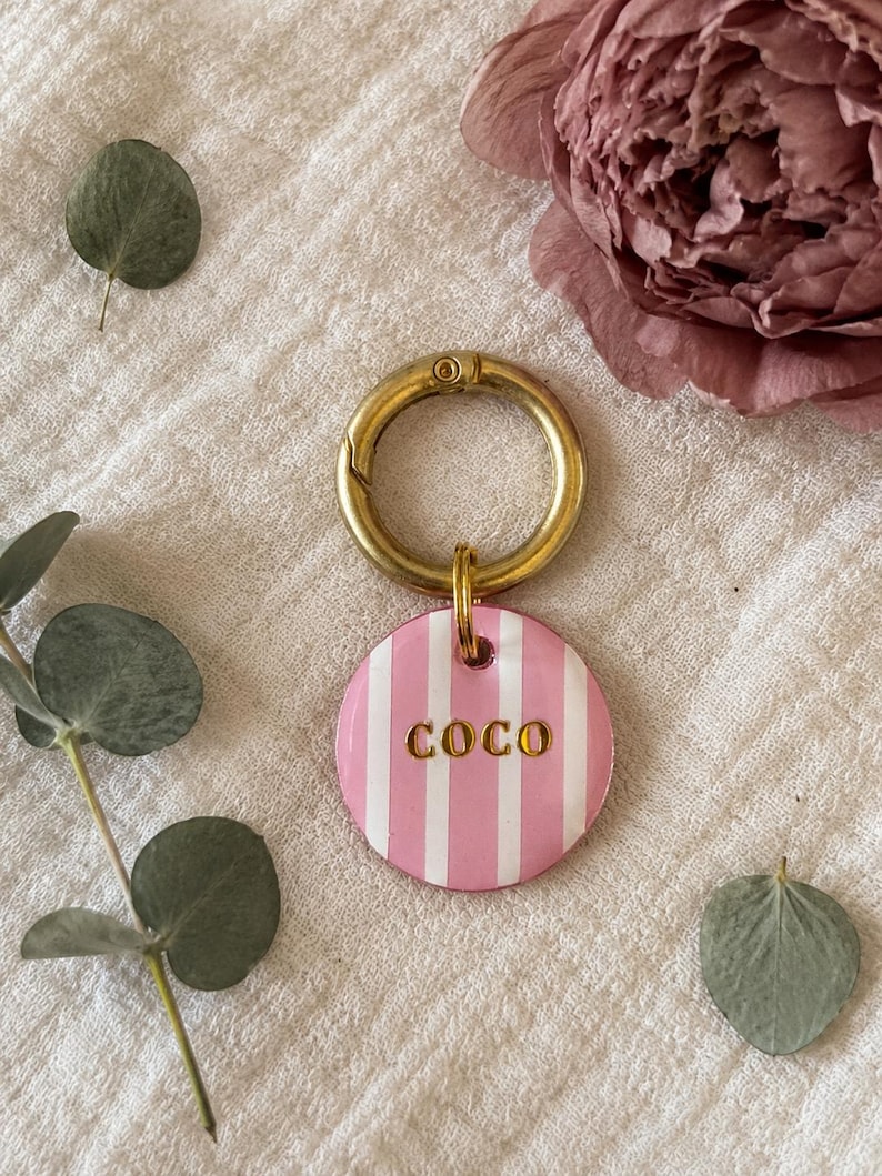 May include: A round pet tag featuring vertical pink and white stripes with the name COCO embossed in gold lettering. The tag is attached to a circular gold-toned metal keyring, providing a secure attachment point for a collar or keychain.